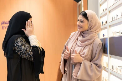 An Emirati woman helps her friend choose the glasses that suit her facial features, the positive impact of shopping on women, choosing suitable sunglasses, the pleasure of shopping from an Emirati mall, the concept of fashion, elegance and interest in one's appearance, two Saudi Arabian Gulf friends wearing an abaya, one of them trying on glasses