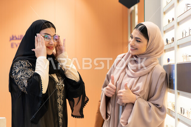 التأثير الإيجابي للتسوق على المرأة، اختيار نظارة شمسيه مناسبة ،متعة الشراء من احدي المولات الاماراتية، مفهوم الموضة والاناقة والاهتمام بالمظهر الخارجي، صديقتان عربيتان خليجيتان سعوديتان ترتديان العباءة يقومان باختيار النظاره الملائمه لملامح الوجه ، امرأة اماراتية تقوم بتجربة النظارات قبل الشراء