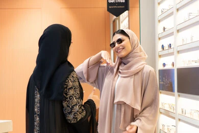 ايماءات الاعجاب والثقة بالنفس ، اختيار نظارة شمسيه مناسبة، احدث صيحات الموضة، صديقتان عربيتان خليجيتان سعوديتان ترتديان العباءة احداهما تقوم بقياس نظاره ، امرأة اماراتية تقوم بتجربة النظارات قبل الشراء، الاهتمام بالمظهر الخارجي،التأثير الإيجابي للتسوق على المرأة