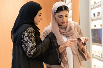 متعة الشراء من احدي المولات الاماراتية، مفهوم الموضة والاناقة والاهتمام بالمظهر الخارجي، امرأة اماراتية تساعد صديقتها فى اختيار النظاره الملائمه لملامح الوجه ، التأثير الإيجابي للتسوق على المرأة، اختيار نظارة شمسيه مناسبة ، صديقتان عربيتان خليجيتان سعوديتان ترتديان العباءة احداهما تقوم بقياس النظارة