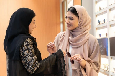 The positive impact of shopping on women, the pleasure of shopping from an Emirati mall, the concept of fashion, elegance and interest in one's appearance, an Emirati woman helping her friend choose glasses that suit her facial features, choosing suitable sunglasses, two Saudi Arabian Gulf friends wearing an abaya, one of them trying on glasses