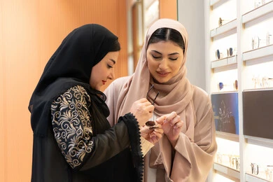مفهوم الموضة والاناقة والاهتمام بالمظهر الخارجي، اختيار نظارة شمسيه مناسبة ، التأثير الإيجابي للتسوق على المرأة، متعة الشراء من احدي المولات الاماراتية، امرأة اماراتية تساعد صديقتها فى اختيار النظاره الملائمه لملامح الوجه ، صديقتان عربيتان خليجيتان سعوديتان ترتديان العباءة احداهما تقوم بقياس النظارة