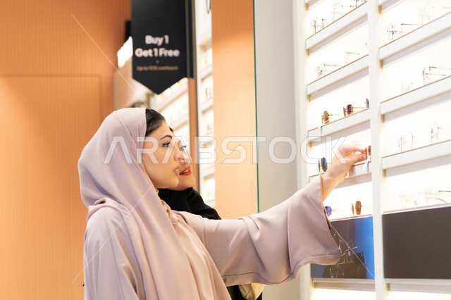 اكسسوارات نسائية عصرية، اختيار نظارة شمسيه مناسبة،صديقتان عربيتان خليجيتان سعوديتان ترتديان العباءة احداهما تقوم بقياس نظاره ورؤيتها على المرآة، امرأة اماراتية تقوم بتجربة النظارات قبل الشراء،  الاهتمام بالمظهر الخارجي،التأثير الإيجابي للتسوق على المرأة