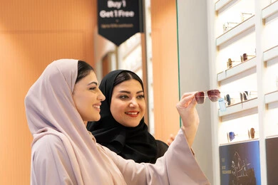 صديقتان عربيتان خليجيتان سعوديتان ترتديان العباءة احداهما تقوم بقياس نظاره ورؤيتها على المرآة، امرأة اماراتية تقوم بتجربة النظارات قبل الشراء،  الاهتمام بالمظهر الخارجي،التأثير الإيجابي للتسوق على المرأة، اكسسوارات نسائية عصرية، اختيار نظارة شمسيه مناسبة