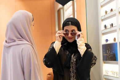 التأثير الإيجابي للتسوق على المرأة،اكسسوارات نسائية عصرية، اختيار نظارة شمسيه مناسبة، الاهتمام بالمظهر الخارجي، امرأة اماراتية تقوم بتجربة النظارات قبل الشراء، صديقتان عربيتان خليجيتان سعوديتان ترتديان العباءة احداهما تقوم بقياس نظاره ورؤيتها على المرآة