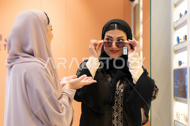 Modern women's accessories, choosing the right sunglasses, the positive impact of shopping on women, taking care of one's appearance, an Emirati woman trying on glasses before buying, two Saudi Arabian Gulf friends wearing an abaya, one of them trying on her glasses and examining them in the mirror.