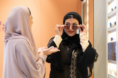اكسسوارات نسائية عصرية، اختيار نظارة شمسيه مناسبة، التأثير الإيجابي للتسوق على المرأة، الاهتمام بالمظهر الخارجي، امرأة اماراتية تقوم بتجربة النظارات قبل الشراء، صديقتان عربيتان خليجيتان سعوديتان ترتديان العباءة احداهما تقوم بقياس نظاره ورؤيتها على المرآة