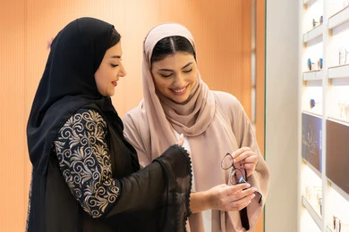 التأثير الإيجابي للتسوق على المرأة ،متعة الشراء من احدي المولات الاماراتية، صورة مقربة لصديقتان عربيتان خليجيتان سعوديتان ترتديان العباءة احداهما تقوم بقياس النظارة ، امرأة اماراتية تساعد صديقتها فى اختيار النظاره الملائمه لملامح الوجه ، الاناقة والاهتمام بالمظهر الخارجي، اختيار نظارة طبية مناسبة ، مفهوم ضعف البصر