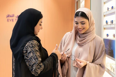 Close-up of two Saudi Arabian Gulf friends wearing an abaya, one of them trying on glasses, an Emirati woman helping her friend choose glasses that suit her facial features, elegance and interest in appearance, choosing suitable medical glasses, the concept of poor vision, the positive impact of shopping on women, the pleasure of shopping in one of the Emirati malls