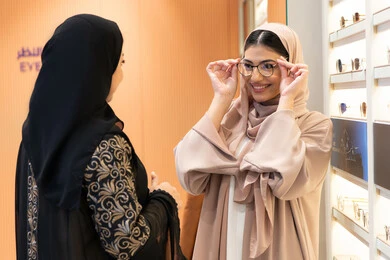 An Emirati woman helps her friend choose the glasses that suit her facial features, elegance and interest in appearance, choosing suitable medical glasses, the concept of poor eyesight, the positive impact of shopping on women, the pleasure of shopping from an Emirati mall, a close-up photo of the hands of two Saudi Arabian Gulf friends wearing an abaya, one of them trying on glasses