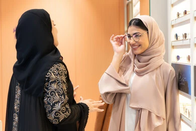 الاناقة والاهتمام بالمظهر الخارجي، اختيار نظارة طبية مناسبة ، مفهوم ضعف البصر ، التأثير الإيجابي للتسوق على المرأة ،متعة الشراء من احدي المولات الاماراتية، صورة مقربة ليد صديقتان عربيتان خليجيتان سعوديتان ترتديان العباءة احداهما تقوم بقياس النظارة ، امرأة اماراتية تساعد صديقتها فى اختيار النظاره الملائمه لملامح الوجه