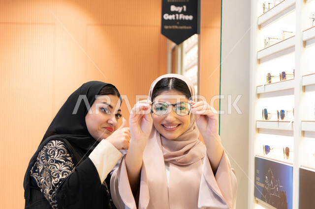 Choosing suitable eyeglasses, the concept of weak vision, elegance and interest in appearance, the positive impact of shopping on women, the pleasure of shopping from one of the Emirati malls, an Emirati woman helping her friend choose the glasses that suit her facial features, two Saudi Arabian Gulf friends wearing the abaya, one of them trying on the glasses