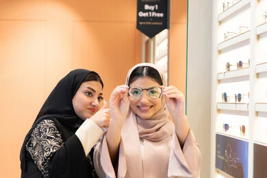 اختيار نظارة طبية مناسبة ، مفهوم ضعف البصر ، الاناقة والاهتمام بالمظهر الخارجي،التأثير الإيجابي للتسوق على المرأة، متعة الشراء من احدي المولات الاماراتية، امرأة اماراتية تساعد صديقتها فى اختيار النظاره الملائمه لملامح الوجه ، صديقتان عربيتان خليجيتان سعوديتان ترتديان العباءة احداهما تقوم بقياس النظارة