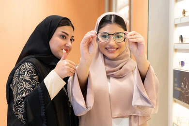 مفهوم ضعف البصر ، الاناقة والاهتمام بالمظهر الخارجي، اختيار نظارة طبية مناسبة ، التأثير الإيجابي للتسوق على المرأة، متعة الشراء من احدي المولات الاماراتية، امرأة اماراتية تساعد صديقتها فى اختيار النظاره الملائمه لملامح الوجه ، صديقتان عربيتان خليجيتان سعوديتان ترتديان العباءة احداهما تقوم بقياس النظارة