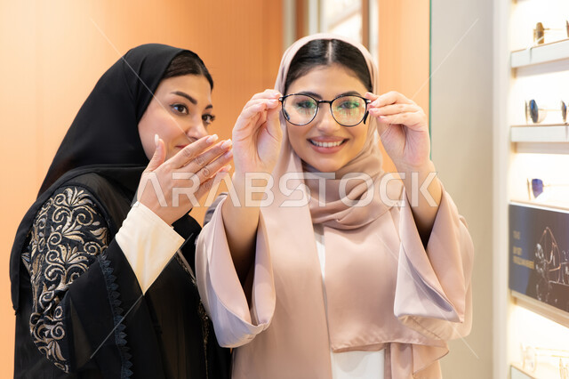 Modern women's accessories, choosing suitable sunglasses, the positive impact of shopping on women, taking care of one's appearance, an Emirati woman trying on glasses before buying, two Saudi Arabian Gulf friends wearing the abaya, one of them trying on her glasses and looking at them in the mirror