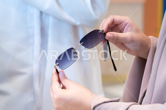 The concept of elegance and attention to one's appearance, shopping and buying sunglasses from a mall. A close-up image of an Emirati or Gulf Arab woman wearing an abaya choosing the right sunglasses in an eyewear store.