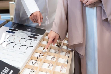 صورة مقربة ليد امراه عربية خليجية اماراتية ترتدي العباءه تقوم باختيار نظاره طبيه، الاهتمام بالمظهر الخارجي، متعة التسويق والشراء ، الحيرة في اختيار نظارة مناسبة، الركن الخاص بالنظارات الطبية ، فحص النظارات الطبيه قبل الشراء