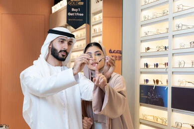 Shopping and buying from malls, paying attention to appearance and elegance, an Emirati woman helping customers in their selection, the section for luxury sunglasses, an Arab Gulf Emirati man wearing the kandura and ghutrah trying on the suitable sunglasses in an Emirati store, discounts and offers.