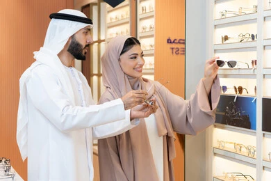 Attention to appearance and elegance, an Emirati woman helping customers in their selection, the section for luxury sunglasses, an Arab Gulf man from the UAE wearing a kandura and ghutrah trying on the suitable sunglasses in an Emirati store, shopping and purchasing from malls, discounts and offers.