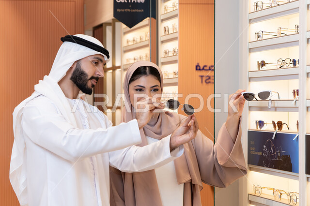 An Emirati woman helps customers in choosing, the section for luxury sunglasses, attention to appearance and elegance, an Arab Gulf Emirati man wearing the kandura and ghutrah is trying on the suitable sunglasses in an Emirati store, shopping and buying from malls, discounts and offers.