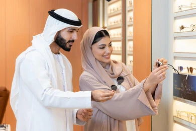 The section for luxury sunglasses, attention to appearance and elegance, an Arab Gulf Emirati man wearing a kandura and ghutrah trying on the suitable sunglasses in an Emirati store, shopping and purchasing from malls, an Emirati woman assisting customers, discounts and offers.