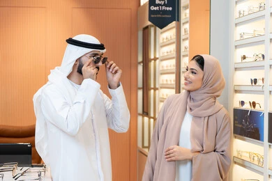 Attention to appearance and elegance, an Arab Gulf Emirati man wearing a kandura and ghutrah is trying on the suitable sunglasses in an Emirati store, shopping and buying from malls, an Emirati woman assisting customers, the section for luxury sunglasses, discounts and offers.