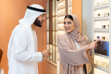 A Gulf Arab Emirati man wearing a kandura and ghutrah is trying on the suitable sunglasses in an Emirati store, shopping and buying from malls, an Emirati woman assisting customers, the section for luxury sunglasses, attention to appearance and elegance, discounts and offers.