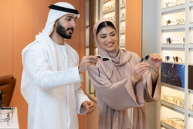 Shopping and buying from malls, an Emirati woman helping customers, the section for luxury sunglasses, attention to appearance and elegance, an Arab Gulf Emirati man wearing the kandura and ghutrah trying on the suitable sunglasses in an Emirati store, discounts and offers.