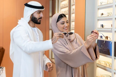 An Emirati woman helps customers in the section for luxury sunglasses, focusing on appearance and elegance. An Arab Gulf Emirati man wearing a kandura and ghutrah is trying on the suitable sunglasses in an Emirati store. There are discounts, promotions, and offers for shopping and buying sunglasses from malls.