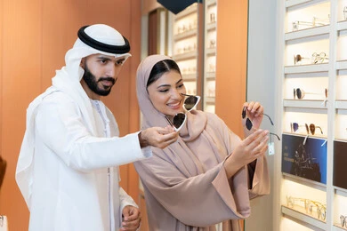 The section dedicated to luxury sunglasses, focusing on appearance and elegance, an Arab Gulf Emirati man wearing a kandura and ghutrah is trying on the suitable sunglasses in an Emirati store, discounts and promotions, shopping and buying sunglasses from malls.