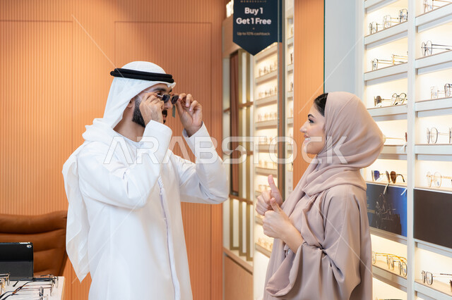 الركن الخاص بالنظارات الشمسيه الفخمه، رجل عربي خليجي اماراتي يرتدي الكندورة والغترة يقوم بقياس النظارة الشمسيه المناسبة في محل نظارات، الاهتمام بالمظهر الخارجي، تخفيضات وخصومات وعروض ، التسوق وشراء النظارات الشمسيه من المولات
