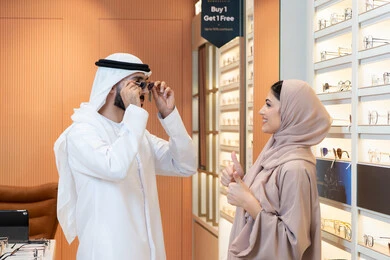 The section for luxury sunglasses, an Arab Gulf Emirati man wearing a kandura and ghutrah is trying on the suitable sunglasses in an eyewear shop, focusing on appearance, discounts and promotions, shopping and buying sunglasses from malls.