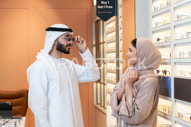 التسوق وشراء النظارات الشمسيه من المولات، الركن الخاص بالنظارات الشمسيه الفخمه، رجل عربي خليجي اماراتي يرتدي الكندورة والغترة يقوم بقياس النظارة الشمسيه المناسبة في محل نظارات، الاهتمام بالمظهر الخارجي، تخفيضات وخصومات وعروض