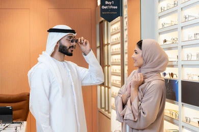 Shopping and buying sunglasses from malls, the section dedicated to luxury sunglasses, an Arab Gulf Emirati man wearing a kandura and ghutrah is trying on the suitable sunglasses in an eyewear store, attention to appearance, discounts and promotions.