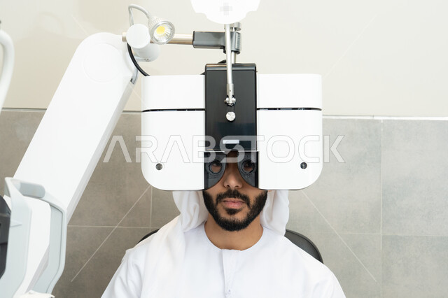 استخدام الأجهزة الحديثة المتطورة، تشخيص وتحديد المشاكل البصرية، تقييم مدى الرؤية والكشف عن الامراض العينية، مفهوم الخدمات والرعاية الصحية في المستشفيات والمراكز،رجل عربي خليجي اماراتي يرتدي الزي التقليدي امام جهاز فحص العين للتأكد من صحة البصر