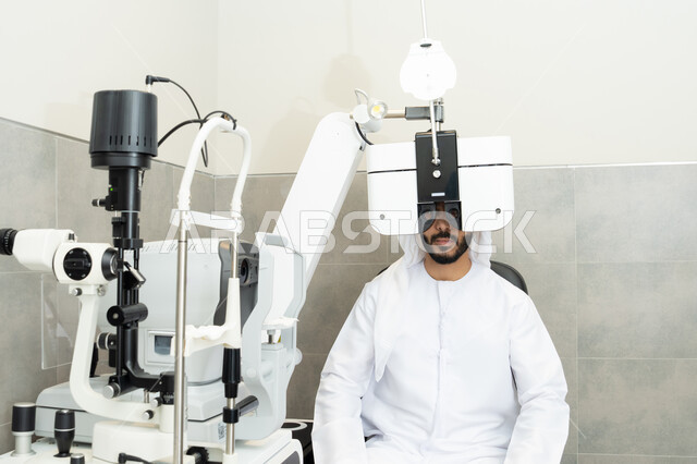 تشخيص وتحديد المشاكل البصرية، استخدام الأجهزة الحديثة المتطورة، تقييم مدى الرؤية والكشف عن الامراض العينية، مفهوم الخدمات والرعاية الصحية في المستشفيات والمراكز،رجل عربي خليجي اماراتي يرتدي الزي التقليدي امام جهاز فحص العين للتأكد من صحة البصر