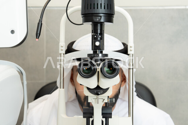 An Arab Gulf Emirati man is examining his eyes with an ophthalmoscope, diagnosing and identifying visual problems, understanding the concept of services and healthcare in hospitals and medical centers, assessing the extent of vision and detecting eye diseases, using advanced modern devices, and ensuring the health of vision.