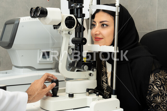 The use of advanced modern devices, a Saudi woman ensures the health of her vision, evaluates the extent of vision and detects eye diseases, diagnoses and identifies visual problems, an Arab Gulf Emirati woman examines her eyes with an ophthalmoscope, the concept of health services and care in hospitals and medical centers.