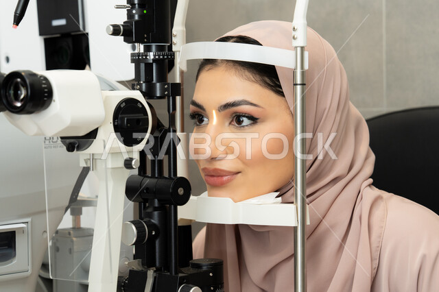 An Arab Gulf Emirati woman is examining her eyes with an ophthalmoscope, a Saudi woman is ensuring the health of her vision, assessing the extent of vision and detecting eye diseases, diagnosing and identifying visual problems, using advanced modern devices, the concept of services and healthcare in hospitals and medical centers.