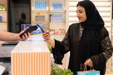 امراة عربيه خليجيه اماراتية ترتدي العباءه السوداء تحمل كيس ورقي ملون بايماءات السعاده و السرور ، سيدة سعودية تقوم بالتسوق والتبضع، إتمام عملية الدفع باستخدام الهاتف المحمول، اقتناء الهدايا من احدي المولات الاماراتيه، تطبيقات الشراء الالكتروني عبر الجوال