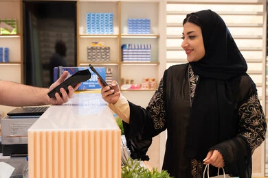 سيدة سعودية تقوم بالتسوق والتبضع، إتمام عملية الدفع باستخدام الهاتف المحمول، اقتناء الهدايا من احدي المولات الاماراتيه، تطبيقات الشراء الالكتروني عبر الجوال ،امراة عربيه خليجيه اماراتية ترتدي العباءه السوداء تحمل كيس ورقي ملون بايماءات السعاده و السرور