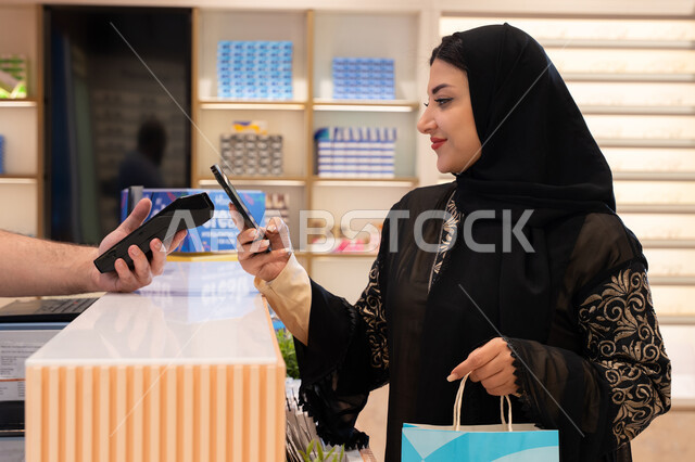 إتمام عملية الدفع باستخدام الهاتف المحمول، اقتناء الهدايا من احدي المولات الاماراتيه، تطبيقات الشراء الالكتروني عبر الجوال ،امراة عربيه خليجيه اماراتية ترتدي العباءه السوداء تحمل كيس ورقي ملون بايماءات السعاده و السرور ، سيدة سعودية تقوم بالتسوق والتبضع