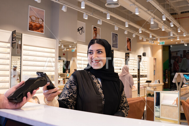 اقتناء الهدايا من احدي المولات الاماراتيه، تطبيقات الشراء الالكتروني عبر الهاتف،امراة عربيه خليجيه اماراتية ترتدي العباءه السوداء تقوم بعملية الدفع باستخدام الهاتف المحمول ،ايماءات السعاده و السرور ، سيدة سعودية تقوم بالتسوق والتبضع