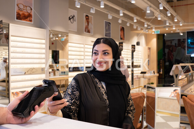 Mobile phone e-commerce applications, purchasing gifts from one of the UAE malls, an Arab Gulf Emirati woman wearing a black abaya is making a payment using her mobile phone, gestures of happiness and joy, a Saudi woman is shopping and buying.