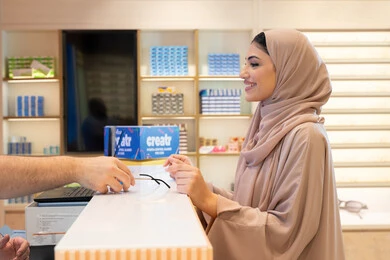 شراء النظارات الخاصة بحماية العين من الاشعه، الاهتمام بالمظهر الخارجي والاناقه والموضة ،  امراه عربيه خليجيه اماراتية محجبه ترتدي العباءه الملونة تقوم بشراء النظارة الشمسيه المناسبة في محل اماراتي بايماءات السعاده و السرور ، سيدة سعودية تقوم بالتسوق والتبضع