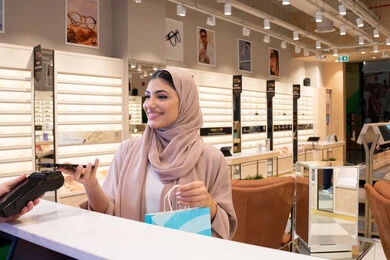 Mobile electronic shopping applications, purchasing gifts from one of the UAE malls, an Arab Gulf Emirati woman wearing a colorful abaya carrying a colorful paper bag with gestures of happiness and joy, a Saudi woman shopping and doing errands, completing the payment process using a mobile phone.