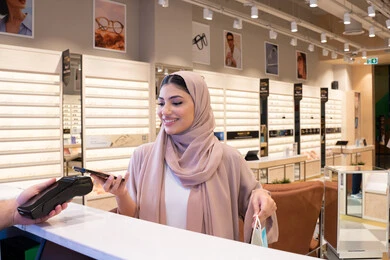 إتمام عملية الدفع باستخدام الهاتف المحمول، تطبيقات الشراء الالكتروني عبر الهاتف، اقتناء الهدايا من احدي المولات الاماراتيه، امراة عربيه خليجيه اماراتية ترتدي العباءه الملونه تحمل كيس ورقي ملون بايماءات السعاده و السرور ، سيدة سعودية تقوم بالتبضع والتسوق
