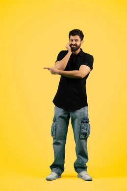 Gestures of boredom and disappointment, concept of elegance and interest in outward appearance, portrait of a smiling young Arab Gulf Emirati man wearing casual clothes standing straight and looking at the camera, Saudi man putting his hand on his face and pointing with his index finger at something, full body, yellow background