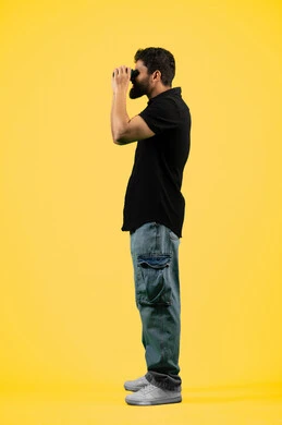 رؤى مستقبلية عند الشباب، البحث عن شيء ما وتوسيع مجال الرؤية، مفهوم الاستكشاف والمغامرة، بورتريه من الجانب لشاب عربي خليجي إماراتي يرتدي الزي الكاجوال يقوم بالنظر من خلال المنظار ثنائي العينين، رجل سعودي يبحث ويستكشف، طول الجسم بالكامل، خلفيه صفراء