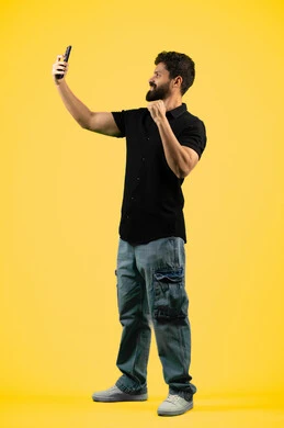 Raising fist in victory gesture, live broadcast on social media, portrait of a young Saudi man in casual clothes taking souvenir photos with a mobile phone, Arab Emirati Gulf man answering a video call, full body image, yellow background